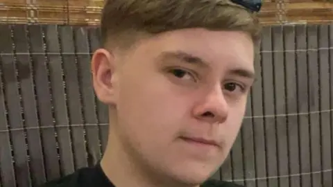 A 15-year-old boy looks at the camera slightly sideways. He has a neutral expression. He wears a black t-shirt.