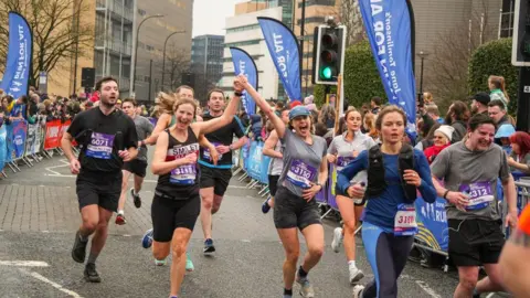 Two women running in the half marathon joining hands and cheering, flanked on either side by supporters behind steel barriers, during the Sheffield Half Marathon last year.