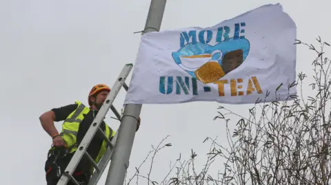Adam Yosef Luke Perry on a ladder attaching one of the flags to a lamppost. He's wearing a high-vis jacket and helmet