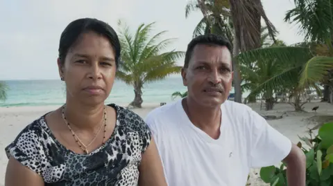 BBC/Andy Alcroft Mr and Mrs Seesahai stare at the camera with solemn faces; they are on a beach in Anguilla, with palm trees and the sea visible in the background