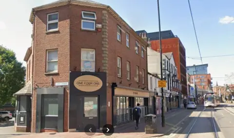 In the foreground is a three-storey red-brick building with a gold coloured oval-shaped sign bearing the words One Four One above a black doorway. Further down the city centre street are a mixture of old and new shops and offices and a tram line on the road. It is a sunny day with a blue sky.