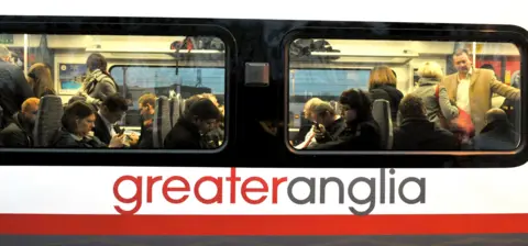 PA Media A close up of a Greater Anglia train carriage. Passengers can be seen inside sitting on the seats and standing in the aisle.