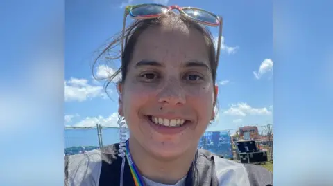 A young woman smiles into the camera. She has dark hair pulled back from her face and sunglasses perched on top of her head. Behind her can be seen sea and blue sky with white fluffy clouds.