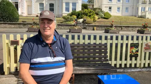 BBC A man in a blue top with white and blue stripes. He is wearing a brown cap. Behind him is a bench. There is a road with flowerpots on the pavement. There are white houses in the background. 