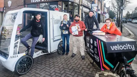 A man on a large white electric cargo bike is stood with four other people, including a man on a smaller black cargo bike.