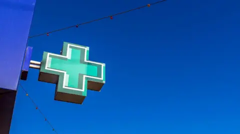 Getty Images A green pharmacy cross sign is mounted on the side of a blue building against a clear blue sky. A string of small coloured lights stretches diagonally across the background. The sign is lit from within and casts a shadow beneath it.