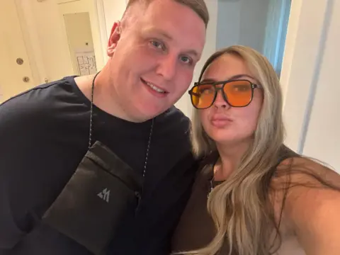 Ellie Mooney Woman with blonde hair and orange lens sunglasses looks at the camera. A blonde man wearing a black top and a cross body bag is standing next to her