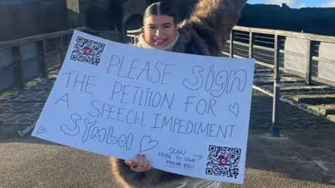 Jessie Yendle Jessie Yendle, who is grinning and holding her campaign poster which says: Please sign the petition for a speech impediment symbol. 