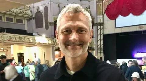 LDRS Si Bellamy, smiling in a hall in Morecambe, with people in the background