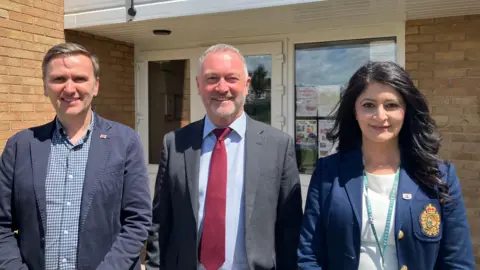 Shariqua Ahmed/ BBC Andrew Pakes, a man in a navy jacket, Steve a man with short grey hair wearing a grey suit and Shabina, a woman in a navy jacket and a white top - all smiling for camera.