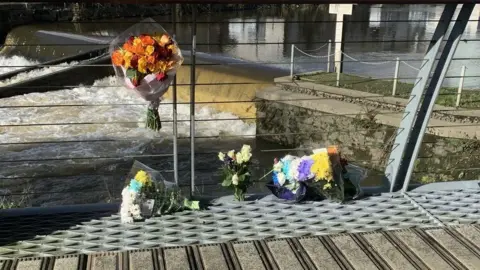 A river, with choppy water and metal fencing in front of it. On the ground, in the forefront, are four bunches of flowers, with  fifth bunch attached to the fencing. qhiddqidqeidzqprw