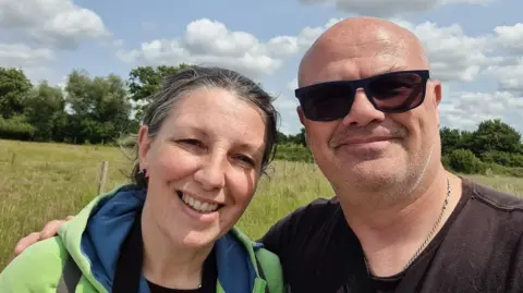 Sompting Wildlife Rescue Gayle and Pete Foggon are pictured out walking. Gayle has tied-back grey hair and is wearing a green and blue jacket, and Pete is bald and is wearing sunglasses and a dark T-shirt. There is a large field and trees in the background.