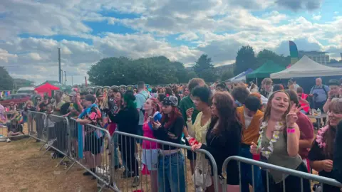 Peterborough Pride A crowd of people are standing behind a metal barrier. There are tents and stalls in the background