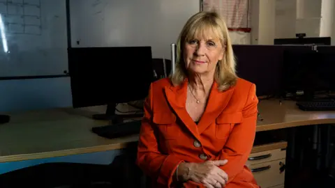 Yeti Television/BBC Cymru Wales Photograph of Penny Roberts, former BBC Wales reporter. She has an orange jacket with brown large buttons in the centre. She has blonde short hair and holds her right arm with her left hand. She is sat on a chair with several computers lined on a desk behind her. 