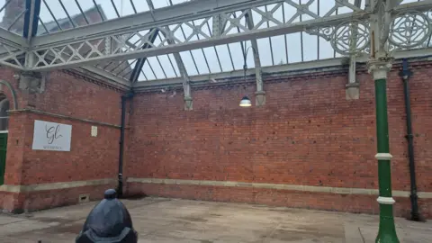 LDRS An empty brick wall at Tynemouth Station where the mural once stood. There are green pillars holding up a glass roof with white iron detailing in a traditional Victorian railway station design. There is a white shop sign on the left hand side. 