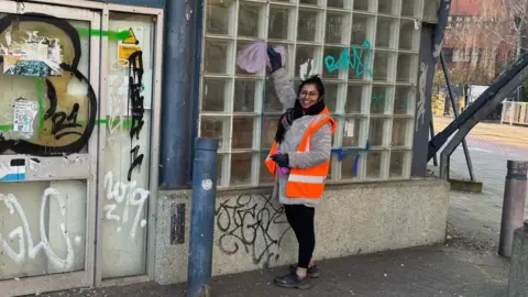 Chrisha Góes Woman in orange high-vis scrubbing a window on an old brick building covered in graffiti