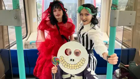 Thomas Copeland / BBC News Three young women in detailed costumes: one in a striped jumpsuit, another in a circular skeleton mask and another in a ruffled red dress.  