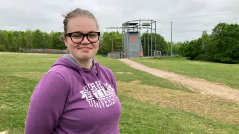 A woman with glasses in a purple hoodie looks at the camera. A tall climbing frame is in the background.