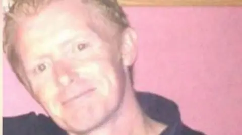 West Yorkshire Police Darren Leach, who has short blonde hair. He is smiling in the photo and wearing a black polo top. He is standing in front of a pink wall.
