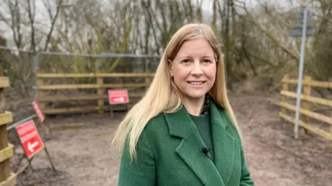 A woman with long blonde hair, green coat and jumper, and small gold hoop earrings, stands at the entrance to the muddy Severn Way path. There are three red and white diversion signs with arrows on behind her. 