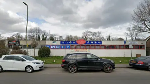 The image shows three cars parked outside an MOT building. Other services can be seen on the signs and the sky is cloudy grey.