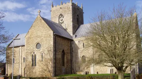 David Justham Exterior view of Stow Minster. The building has a central tower with a clock with arched windows on either side. There is a lawned area in the foreground.