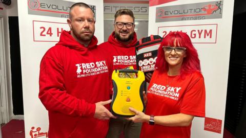 Three people in red tops saying Red Sky Foundation. They are holding CPR kit and a defibrillator. The man to the left is Michael Donkin and he has short hair. The man in the middle is Sergio Petrucci and he has a beard and glasses. The woman to the right is Lisa Loftus. She has long dyed-red hair and glasses