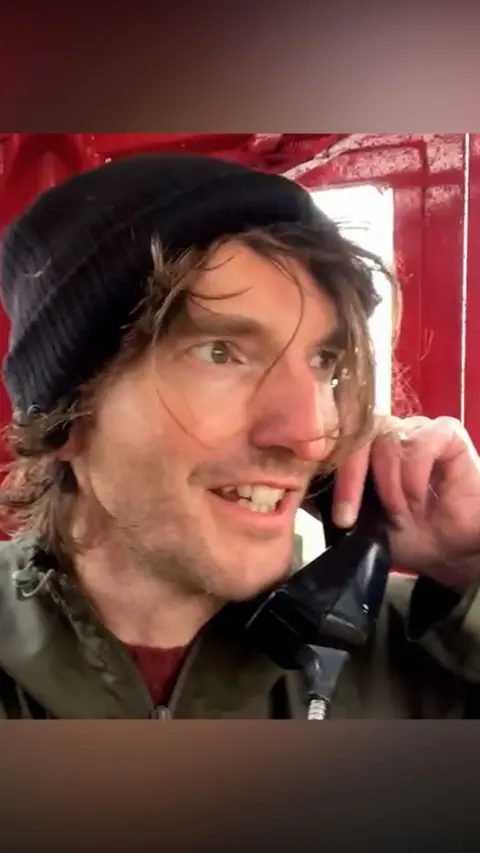 Man with straggly hair in a red phone box