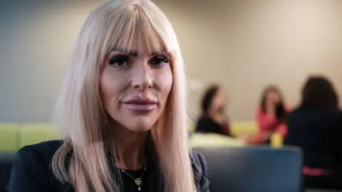 A woman with long, light blonde hair wearing a dark top and a necklace, seated indoors with a blurred background showing three people sitting on green chairs