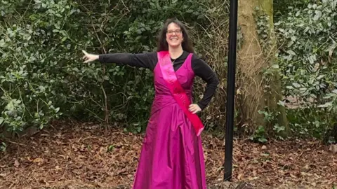 Supplied Jennifer Gane, who has black glasses and long black hair, is wearing a pink ballgown over a black shirt. She is standing in front of trees and is pointing to her right.