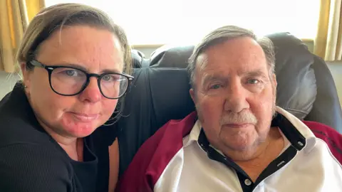 Hayley on the left - she is wearing glasses, has brown hair and a black t-shirt next to her father Russell who is sat in an armchair. He has short grey hair and has a moustache.
