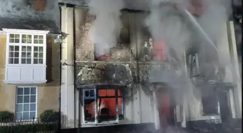 Servicio de Bomberos y Rescate de Dorset y Wiltshire. Un edificio arrasado por el fuego, con ventanas rotas, ceniza por todas partes y humo saliendo de las ventanas. Se puede ver humo rojo del incendio dentro del edificio.