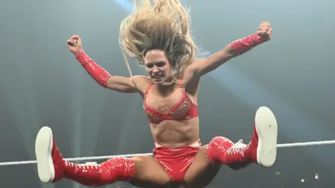Maxxine Dupri is pictured in a really close-up shot, mid-jump in a wrestling ring. She is wearing a red bikini-type outfit with long, red socks and red trainers. She has long blonde hair which is pictured streaming above her head/
