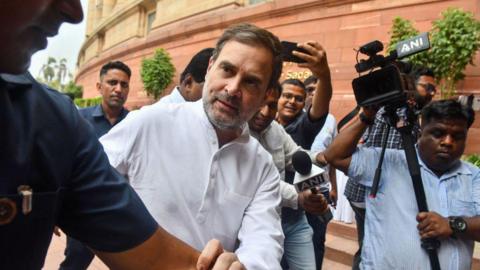 A man wearing a white kurta is seen walking while being closely surrounded by several people, including security personnel and members of the media. The media personnel are holding microphones and cameras, some branded with 