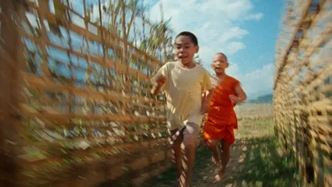 Sandwich Club Productions Blurred imaged suggesting motion of two Laotian children running between two fences with crops behind them