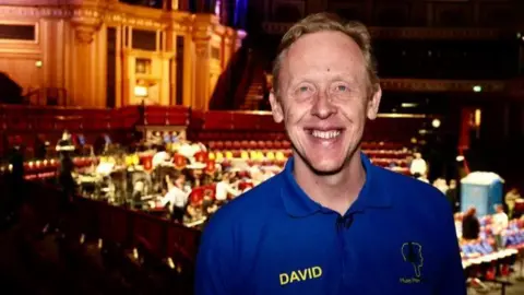 David Stanley is wearing a blue polo shirt and smiling. Behind him a performance is ongoing in a grand looking theatre.