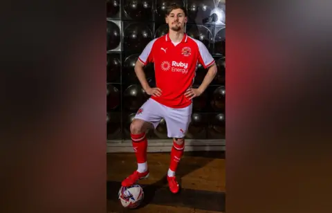 Fleetwood Town FC A man with short dark hair and a goatee beard stands with his hands on his hips and right foot resting on a football. He is wearing a red football shirt with white sleeves, across the front of which is a white Puma sign, the club crest and the sponsor Ruby Energy. He is also wearing white football shorts, red socks pulled up to his knees and red football boots.