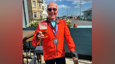 Norman Brown Norman Brown wearing sunglasses smiling at the camera wearing a red waterproof jacket with a medal around his neck holding a pint of beer.
