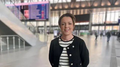 PA Media Liz Kimmins standing inside Grand Central Station in Belfast. She has brown hair tied back, wearing a black and white striped top and black cardigan.
