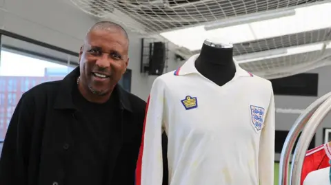 Graham Budd Auctions Viv Anderson, wearing a black jumper and coat, stands smiling beside the England shirt he wore on his debut in 1978. The V-neck jersey is white but slightly faded and has mud stains across the front. An England crest is on the left breast while an Admiral manufacturer's logo is on the opposite side. The long sleeves have red and blue stripes.