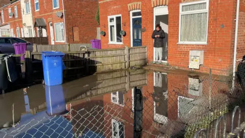 BBC/Paul Murphy A flooded front garden. At the back is a woman wearing a black coat and grey joggers standing on the front doorstep of a red-brick house looking at her mobile phone, which she is holding with both hands