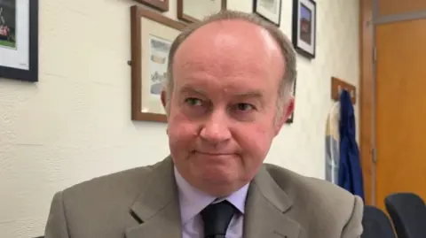 A man with short graying hair. He is wearing a beige jacket, light purple shirt and a dark tie.