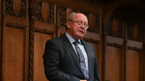 PA Media Jim Allister is bald on top with light hair at the sides. He is speaking in Parliament as he wears a dark suit, light blue shirt and patterened tie