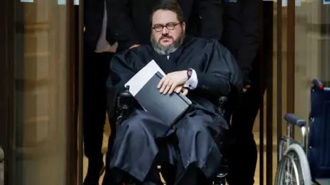 Getty Images Nicholas Rossi in a wheelchair being pushed out of Edinburgh High Court, He is wearing a black robe, glasses and his a grey beard.