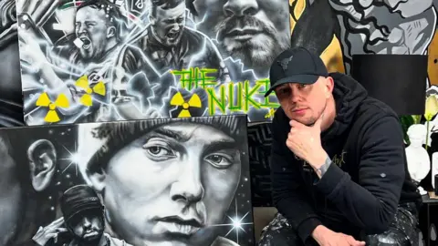 Adam Salisbury Adam Salisbury who has stubble wearing a black baseball cap and black hoodie sits by some of his artwork which including paints of darts star Luke Littler and rapper Eminem. His elbow is resting on his knee and his hands holds his chin. 