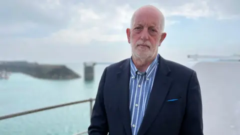 A man wearing a black blazer and a blue and white striped shirt stands outdoors. He is looking at the camera. The sea and land are in the distance behind him. 