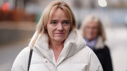 A woman with blonde hair, wearing a white puffer jacket, walks along a street. She is visible from the chest up, with a person walking behind her out of focus. 