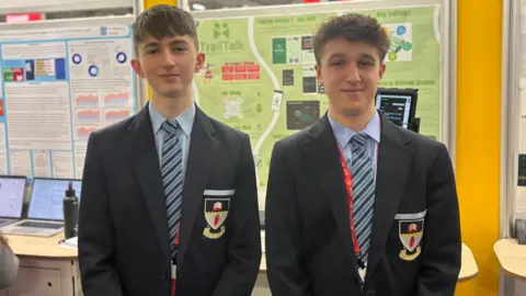 Two teenage boys with brown hair. They're wearing navy blazers with a blue shirt and a red, blue and green stripped tie. A poster on a board behind them says trail talk.