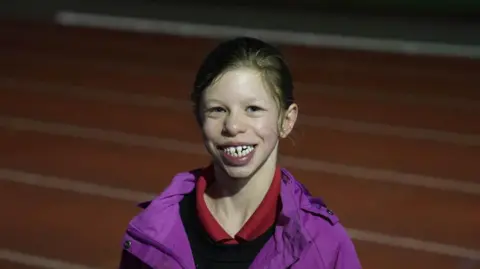 John Fairhall/BBC Fabrienne smiles widely at the camera while standing on the athletics track. She is wearing a purple coat with a black jumper and red polo top on underneath. She has long brown hair which has been tied up behind her head.