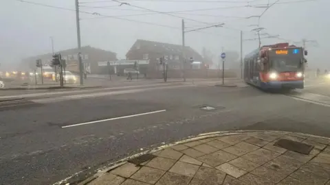 Oli Constable/BBC A misty road in Sheffield with shops in the background and a tram with its lights on turning at a junction.